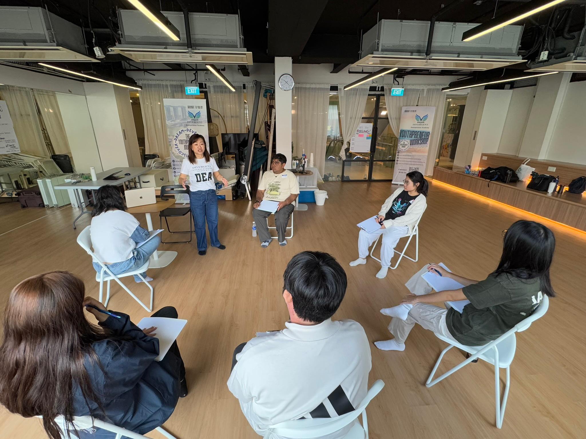 A facilitator in a Divergent Entrepreneur Academy shirt leads a youth workshop circle with participants seated around her